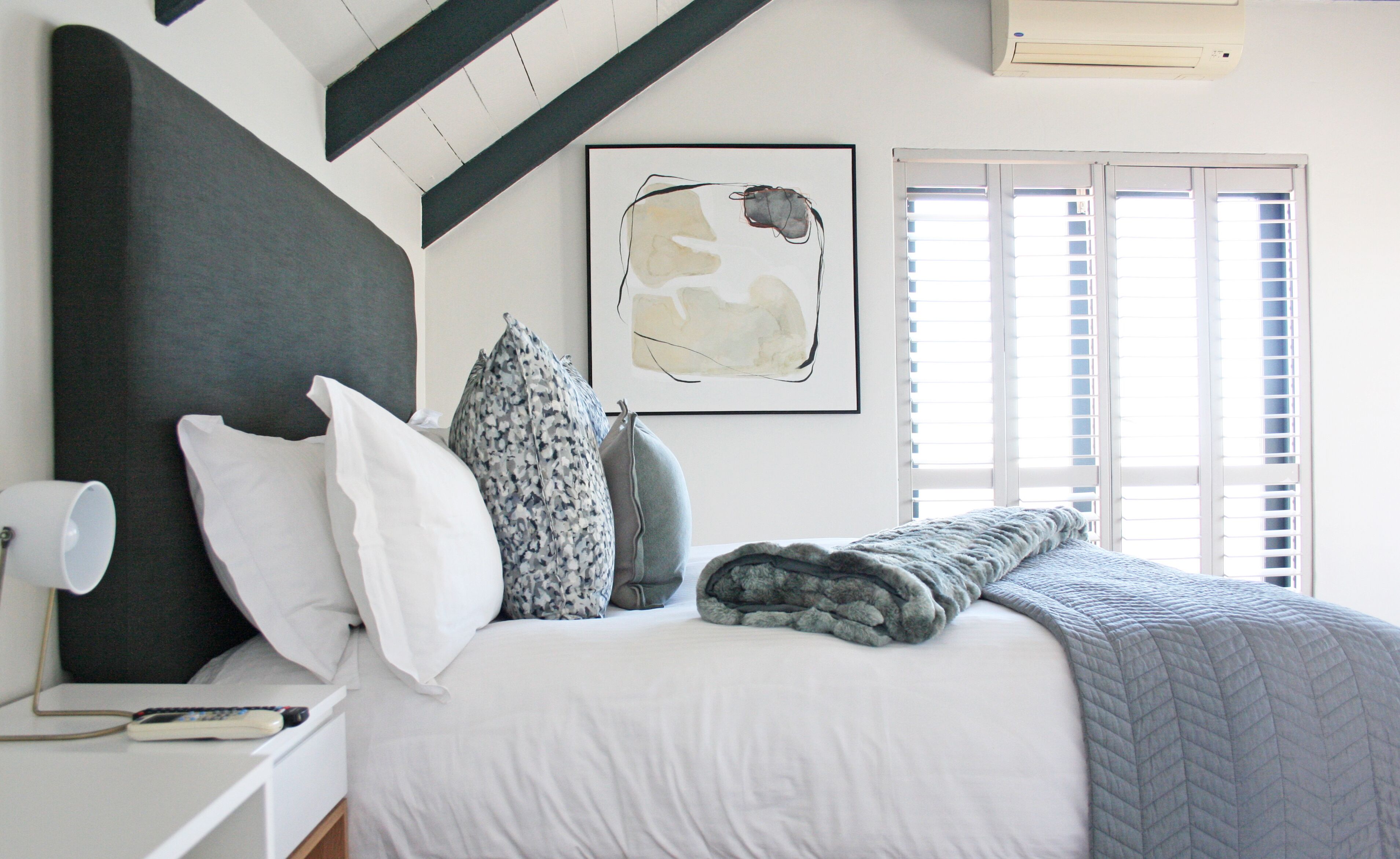 Un dormitorio con cama, almohadas, una colcha, una lámpara de noche, un control remoto y una obra de arte abstracta enmarcada en la pared.