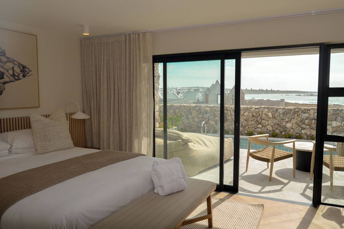 Habitación de hotel con una cama grande, vista al mar y balcón con mobiliario exterior.