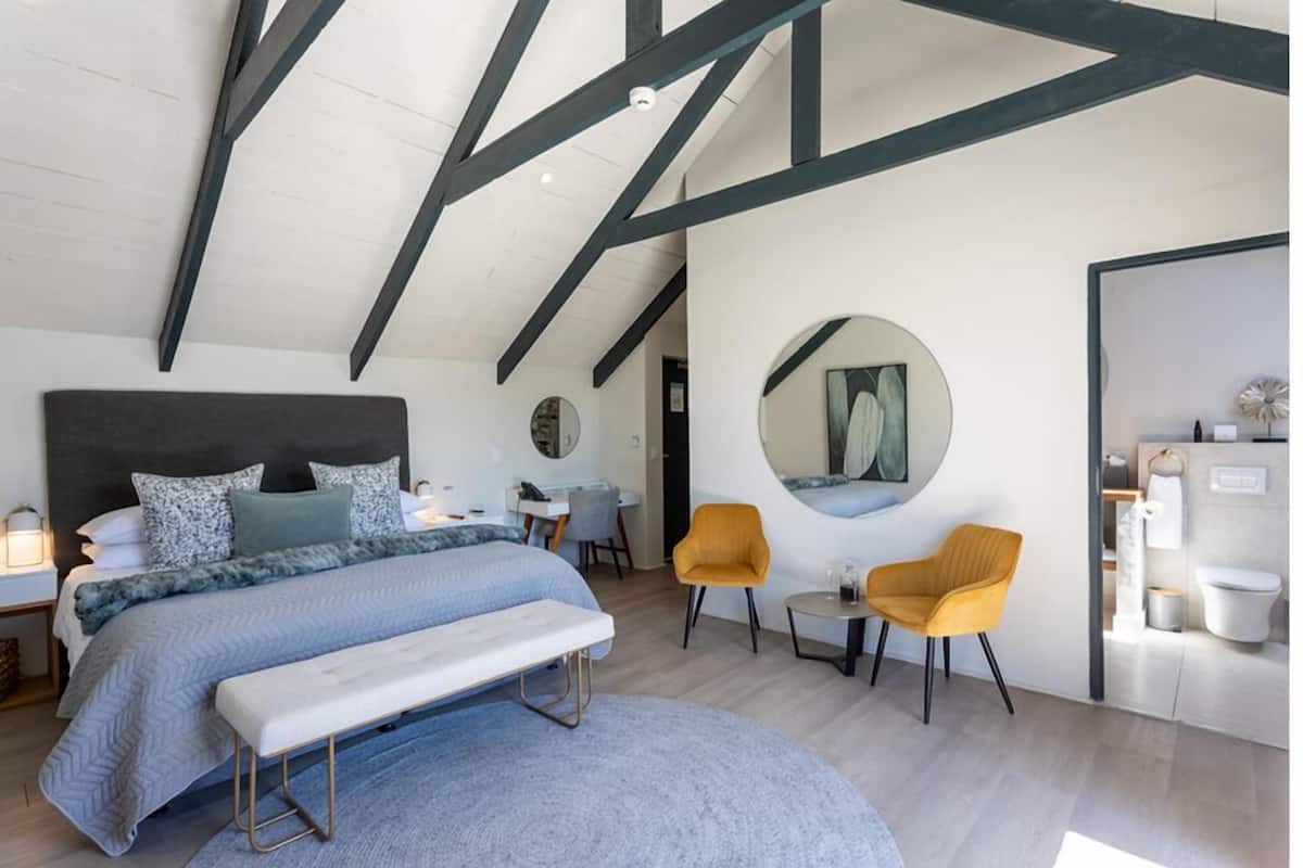 Un dormitorio moderno con una cama grande, un banco, dos sillas, un espejo redondo y un baño visible a través de una puerta abierta.