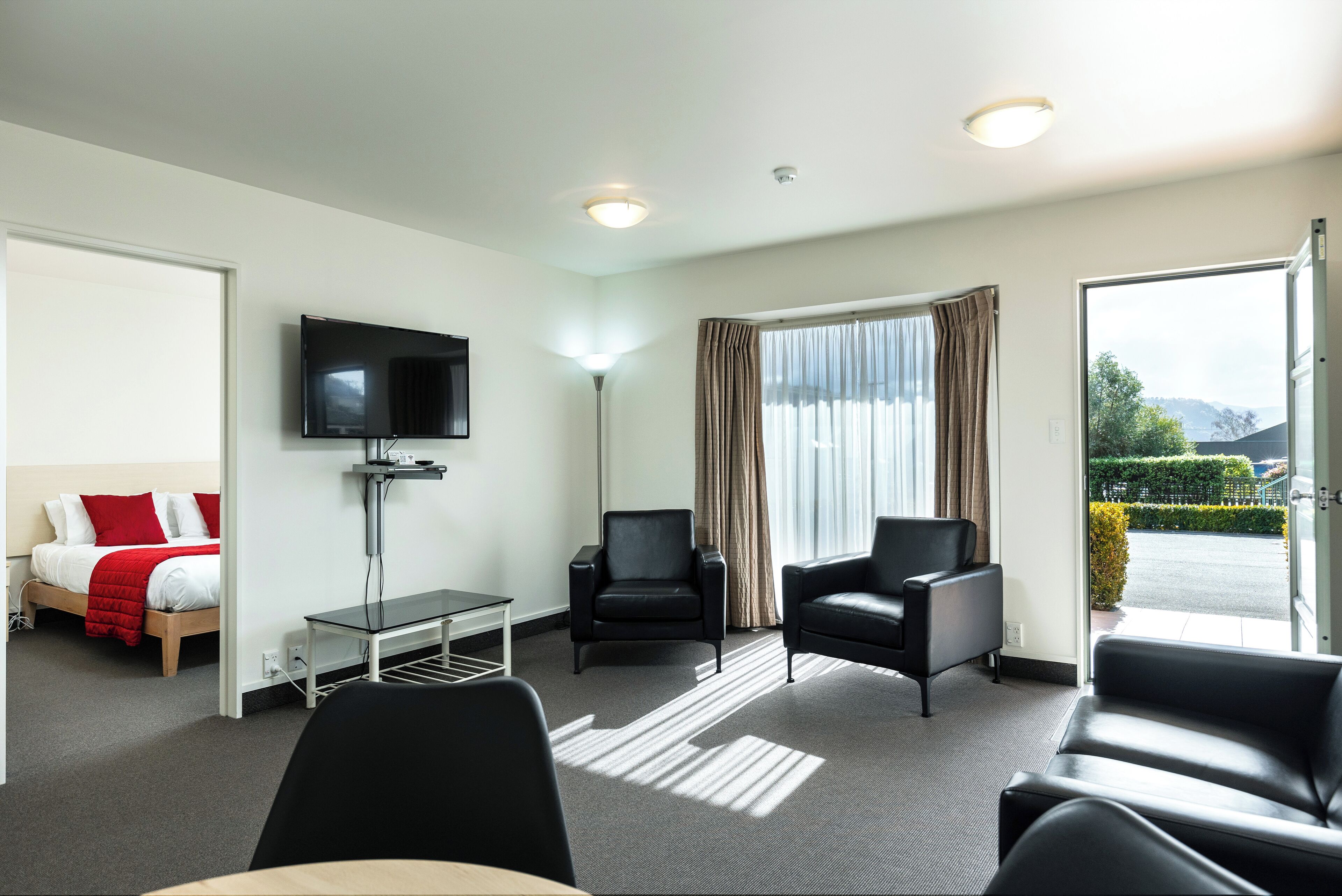 A hotel room with a bed, a TV, a desk, two chairs, and a large window with curtains.