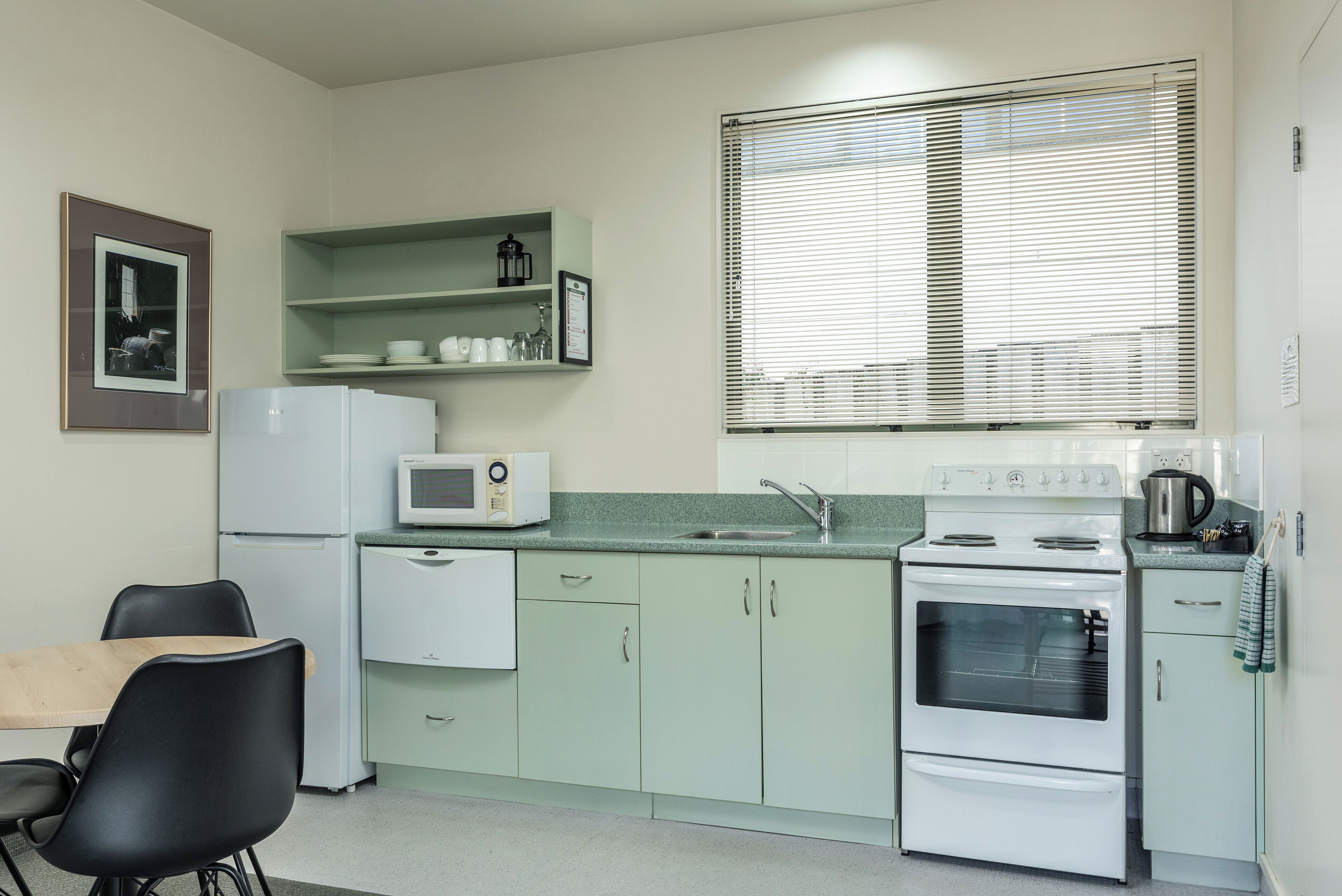 A compact kitchen with a refrigerator, microwave, stove, and sink. There are two chairs and a small table. Shelves above the sink hold dishes and a framed picture.
