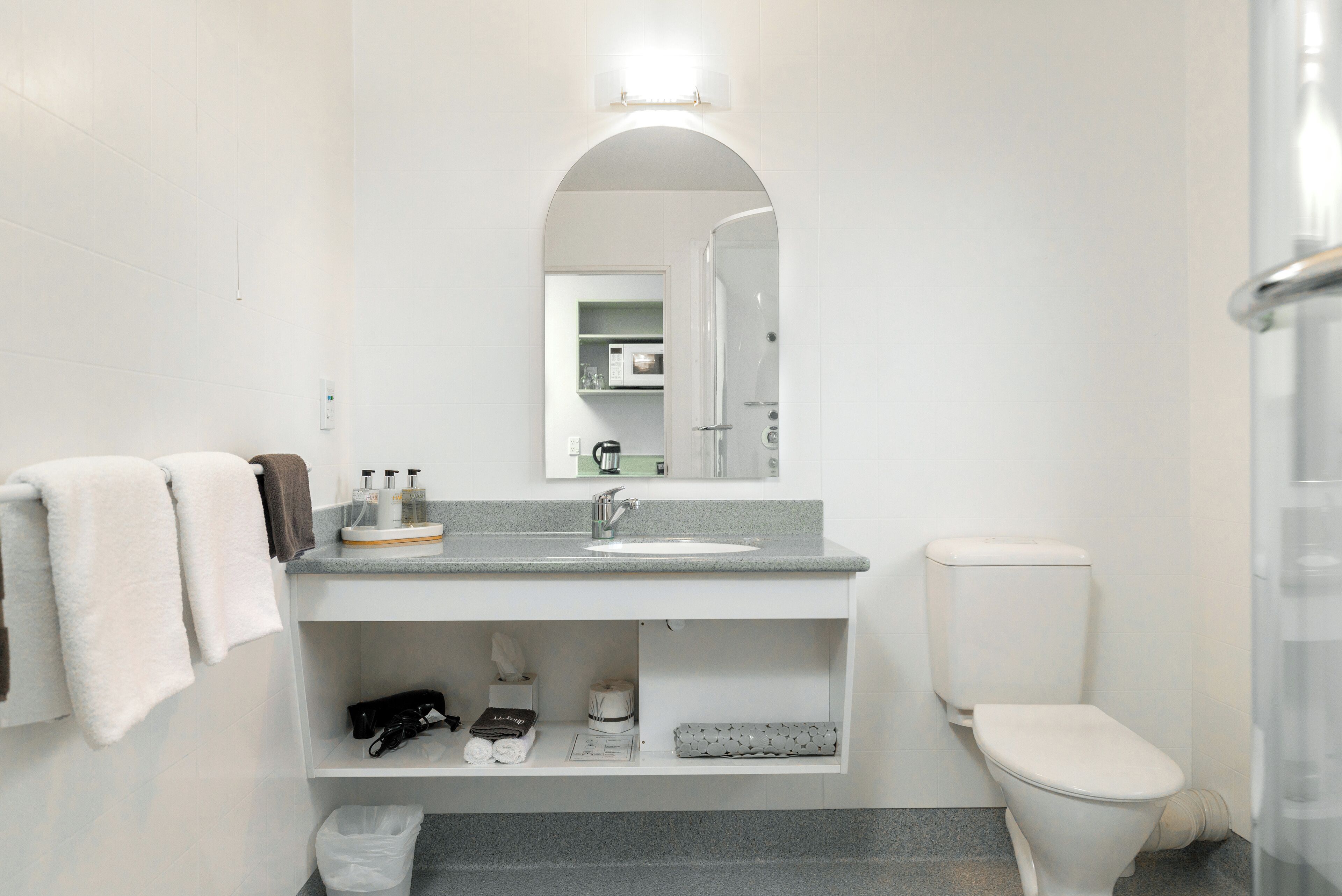 A modern bathroom with a white sink, a toilet, and a mirror above the sink. There are towels hanging on the wall and a trash bin under the sink.