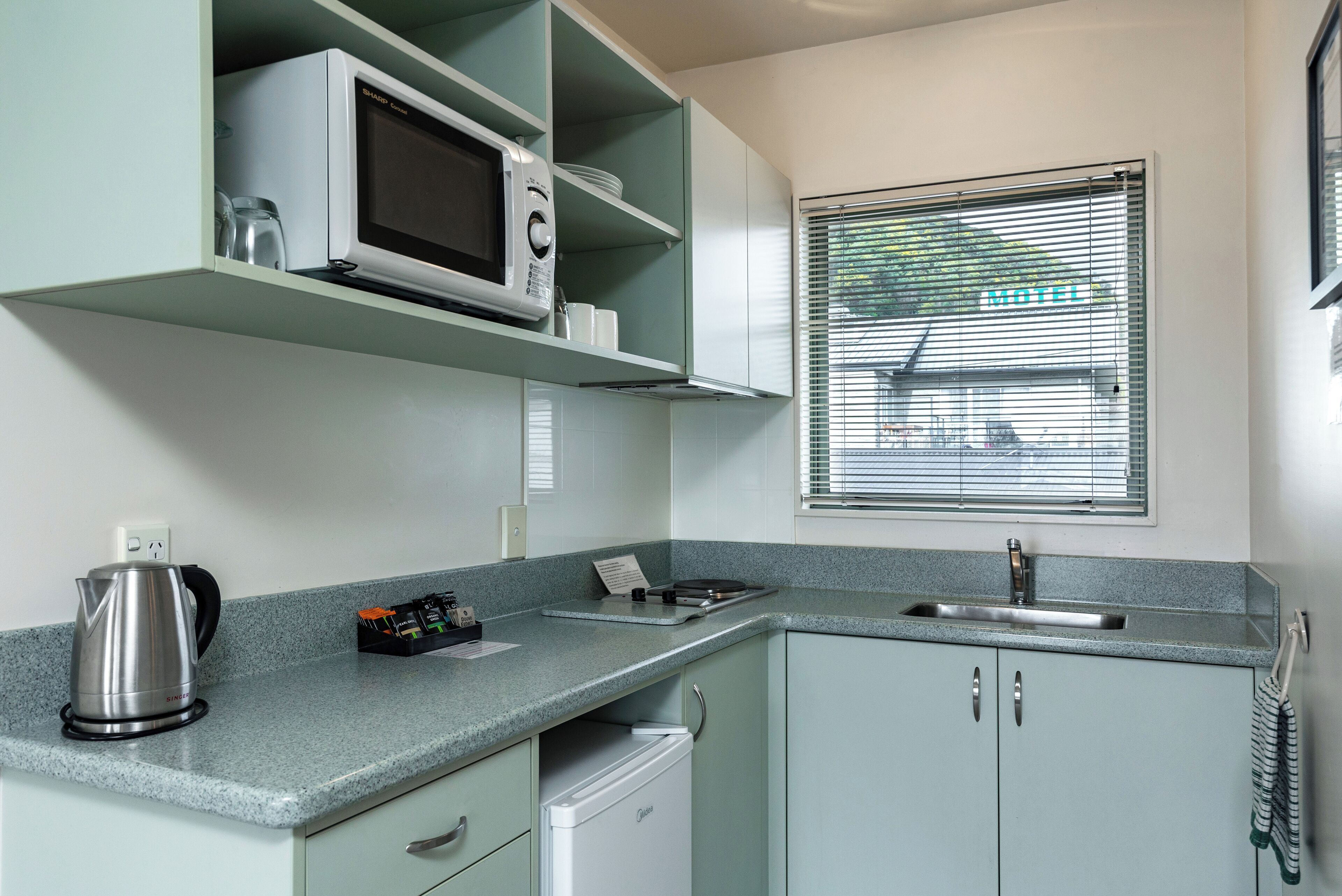 A modern kitchen with a microwave, kettle, and sink. There is a window with a view of a motel sign outside.