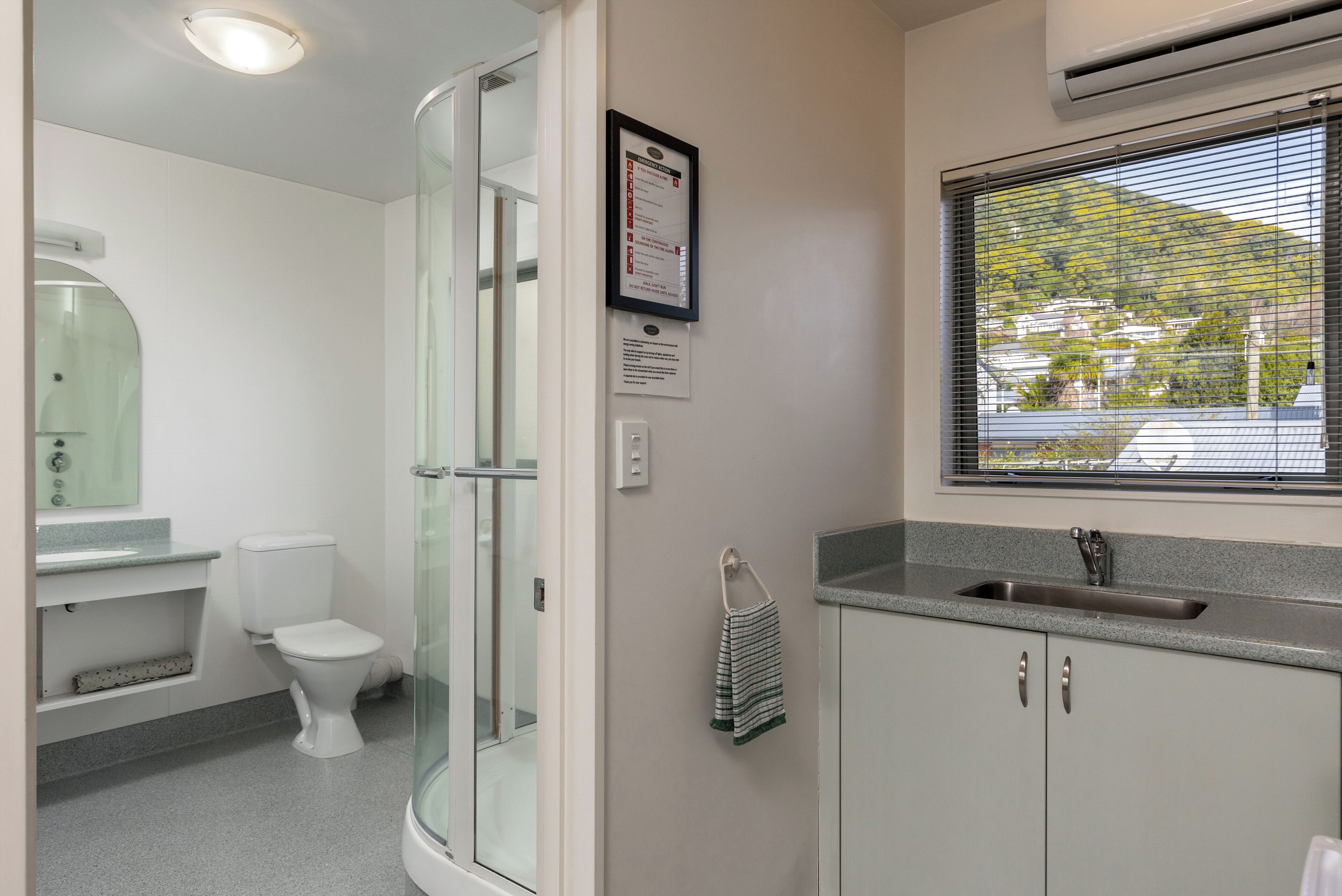 Ein Badezimmer mit Duschkabine aus Glas, Toilette, Waschbecken und einem Fenster darüber sowie einem Handtuchhalter.