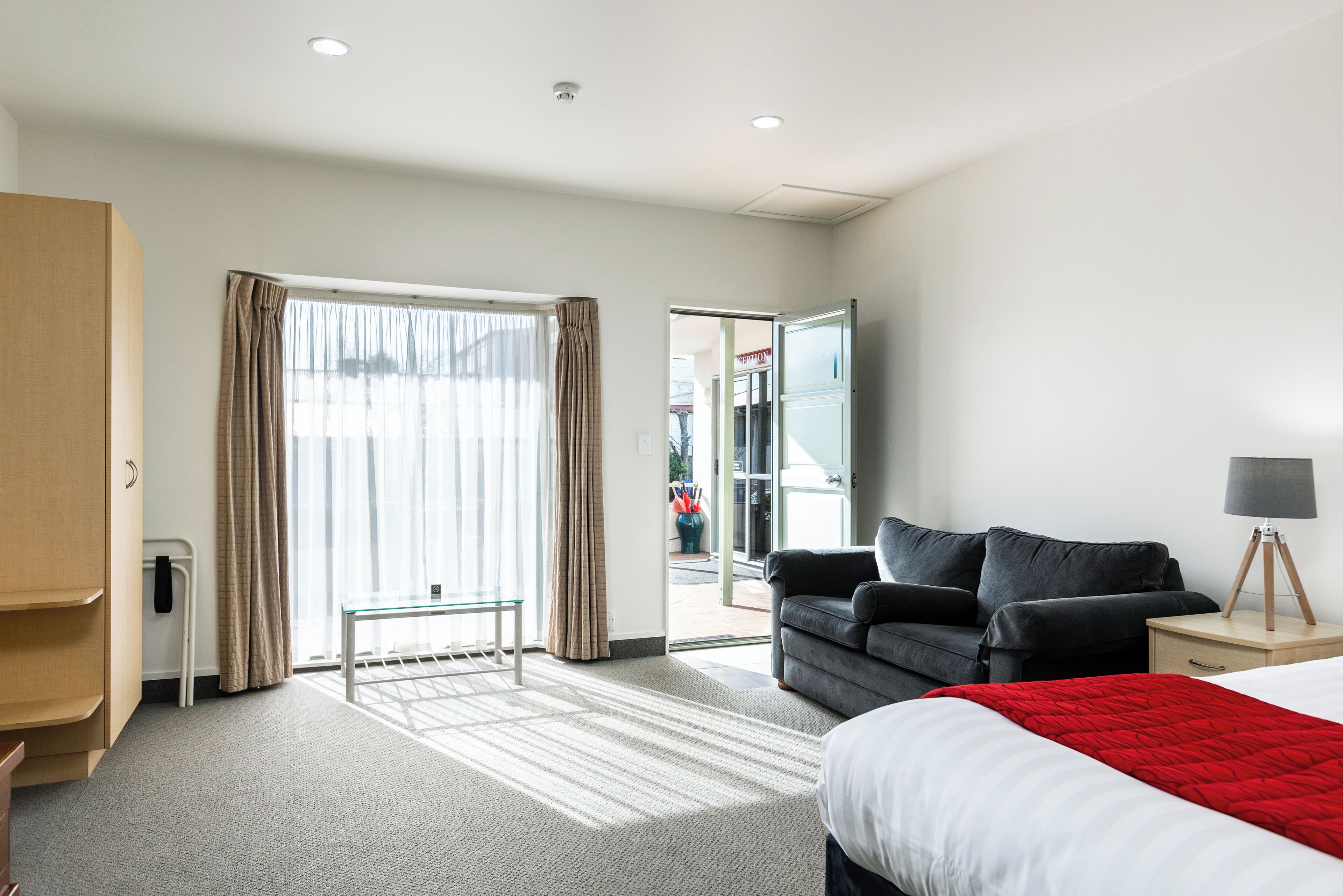 A hotel room with a bed, a sofa, a nightstand, a lamp, a window with curtains, and a door leading to a balcony.