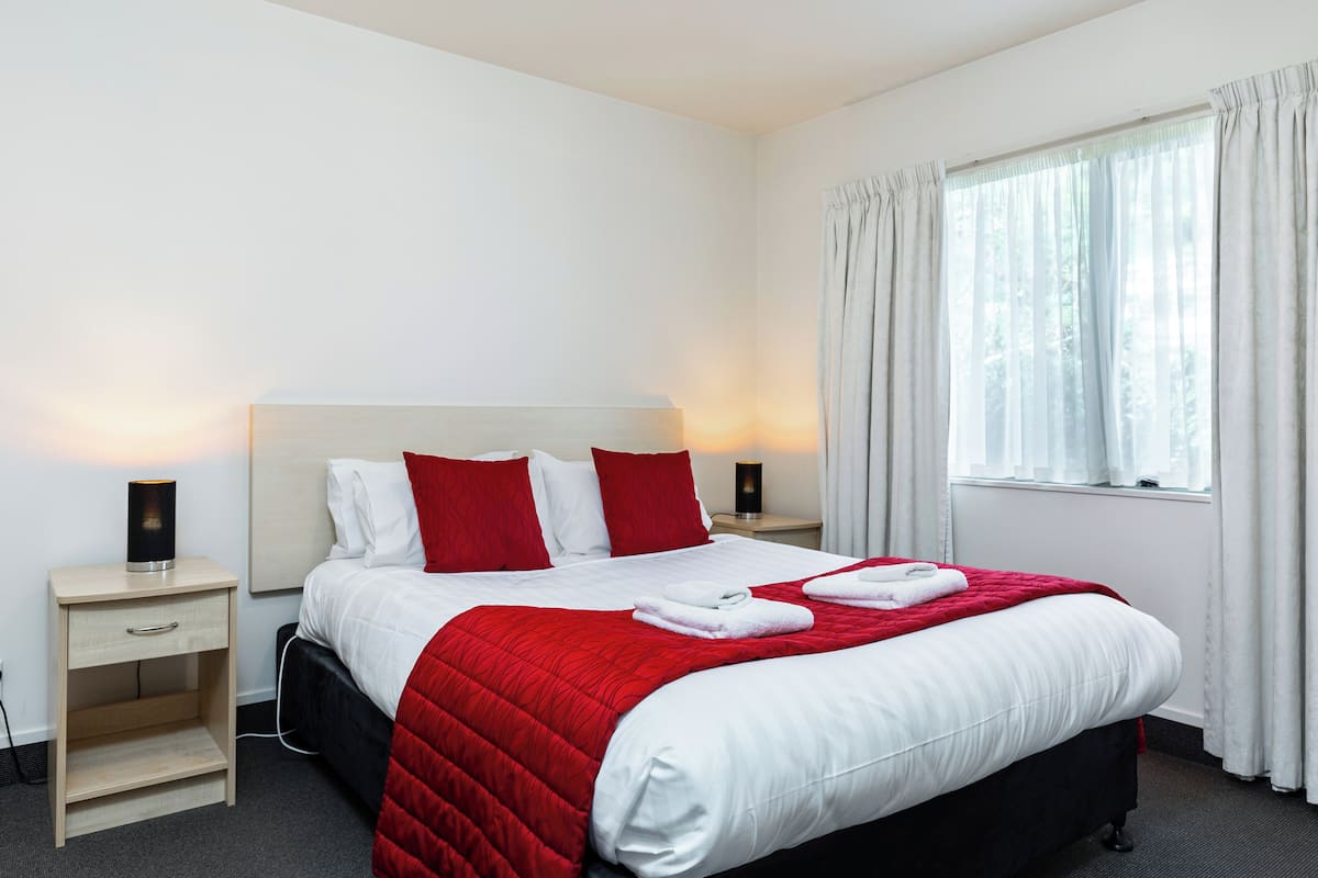 A neatly made bed with a red and white bedspread, two red pillows, and two white towels folded on the bed. There is a bedside table with a lamp on it, and a window with white curtains.