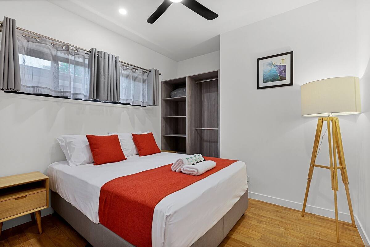 Un lit bien fait, recouvert d’une couverture rouge, une table de chevet, un plancher en bois, une lampe sur pied et une armoire.