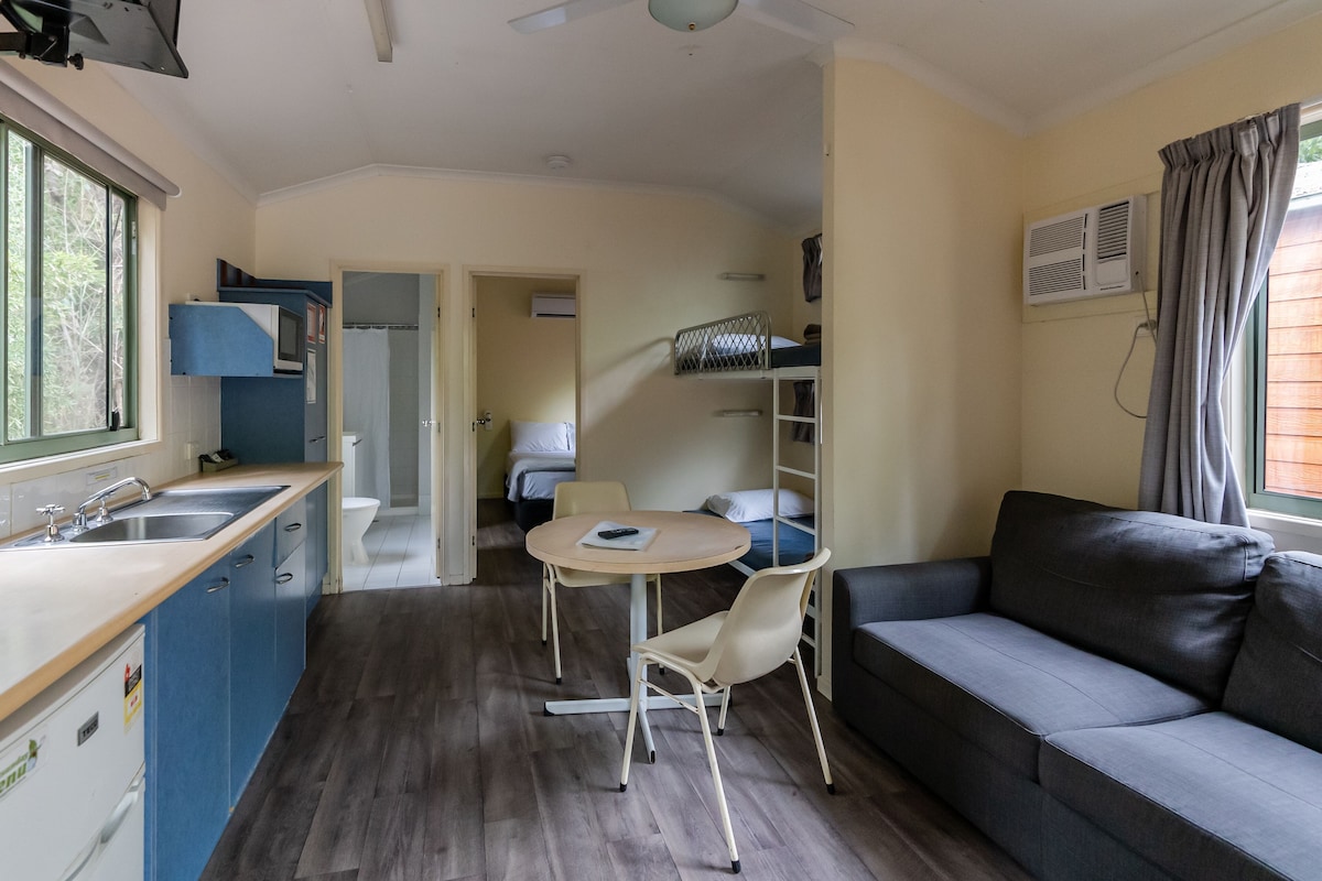 Een compacte kamer met kitchenette, een slaapbank, een eettafel met stoelen en een stapelbed.