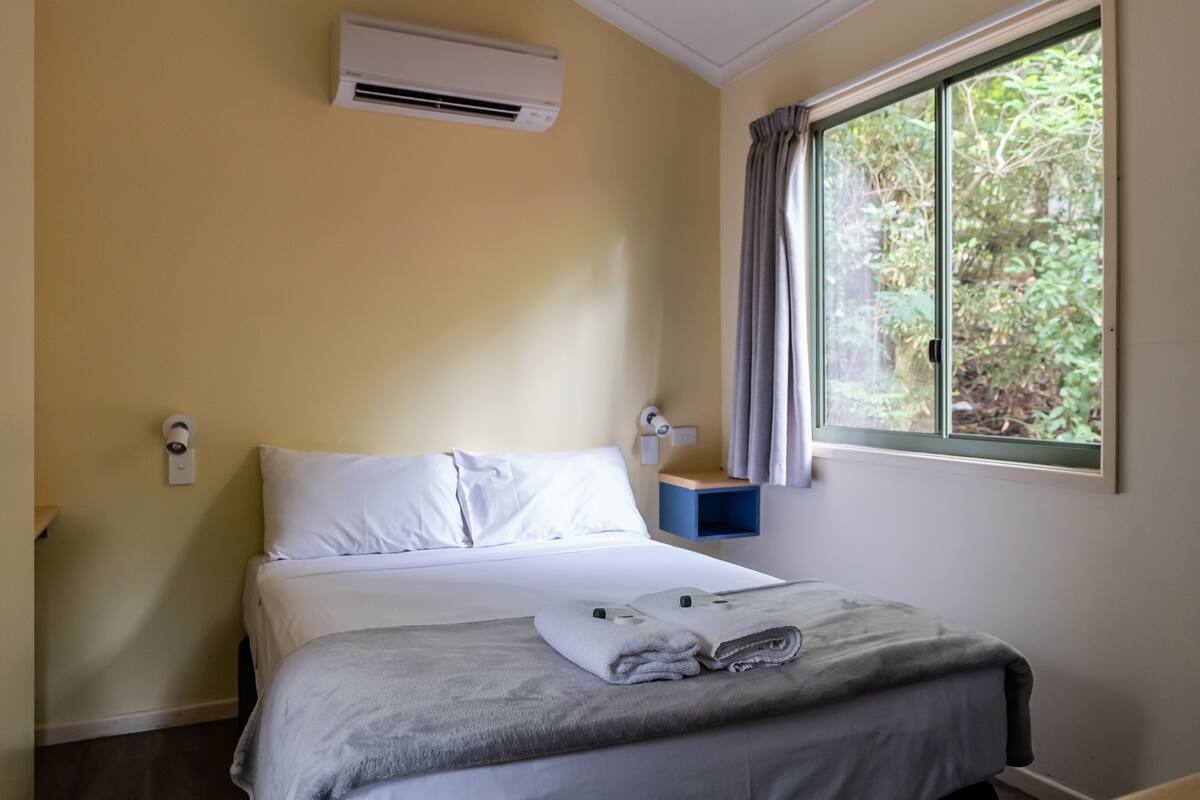 Une chambre d’hôtel avec un lit, un climatiseur, une fenêtre avec des rideaux et une petite table de chevet bleue.