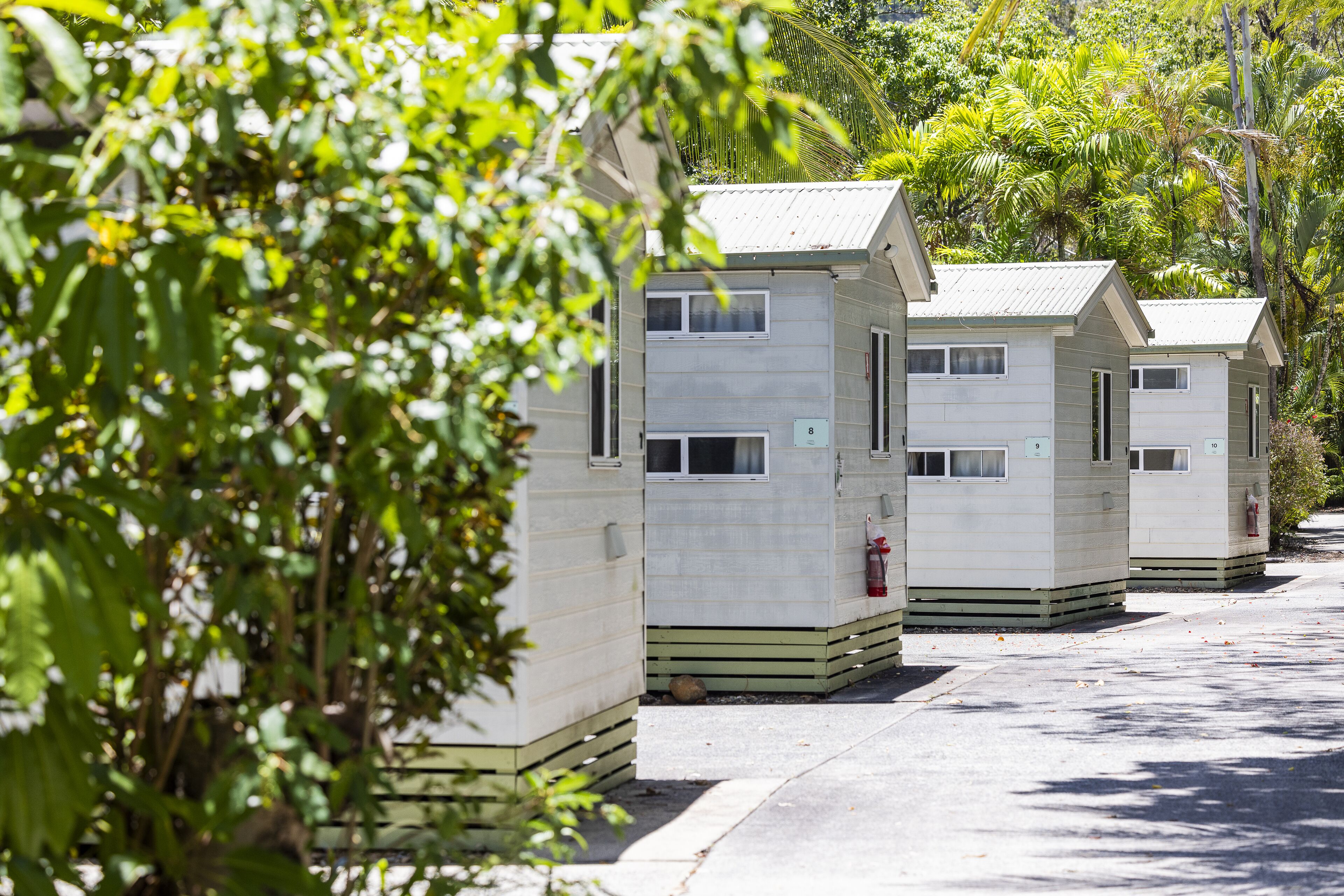 A row of small, single-story cabins with a simple, functional design, each with a small window and a door, situated in a natural setting with trees and greenery around.