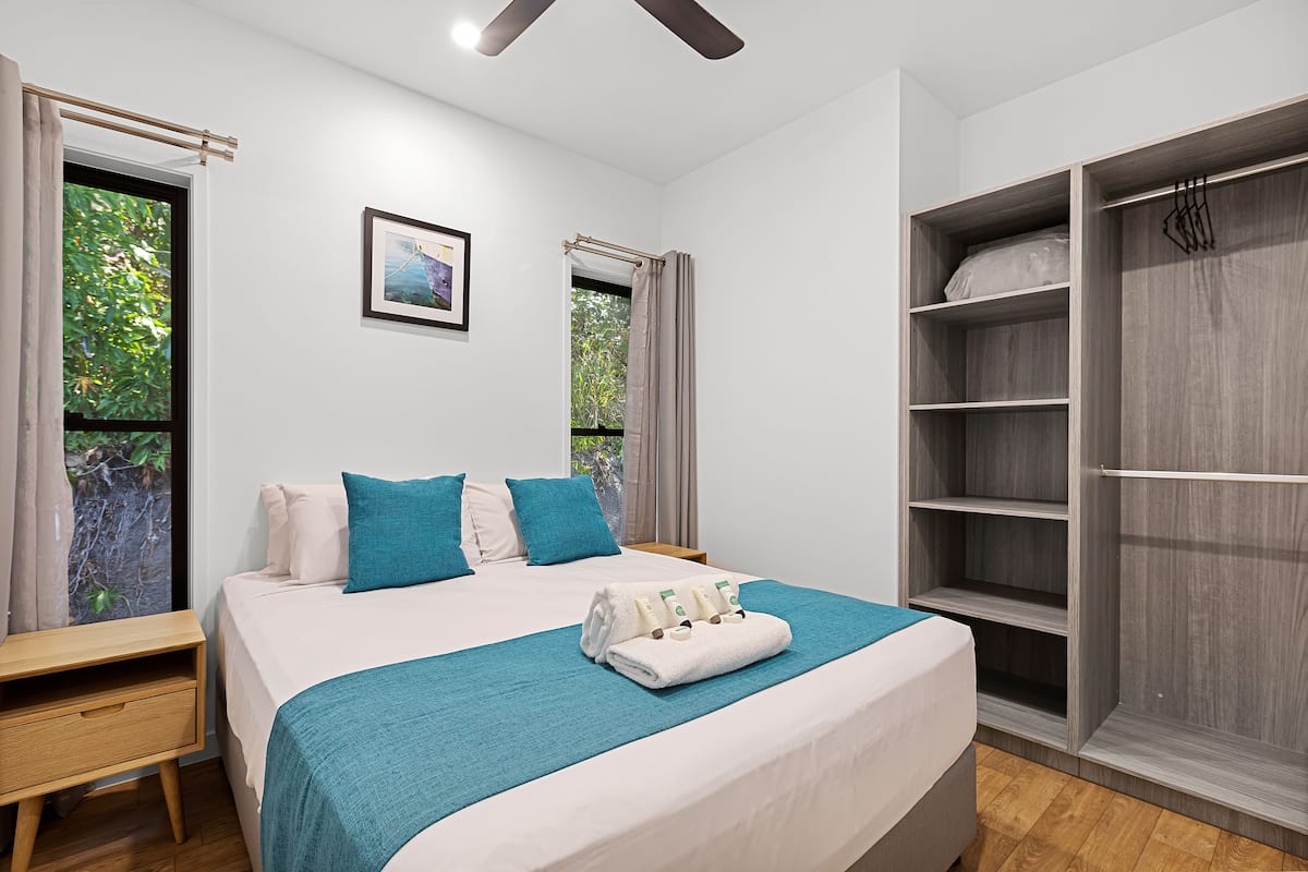 Un lit bien fait, recouvert d’une courtepointe turquoise, avec un oreiller blanc et un oreiller bleu. Une table de chevet en bois, dotée d’un tiroir, se trouve à côté du lit, ainsi qu’une petite table. Une grande armoire à plusieurs étagères est placée à droite du lit.