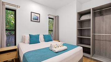 A neatly made bed with a teal bedspread, a white pillow, and a blue pillow. There is a wooden nightstand with a drawer and a small table beside the bed. A large wardrobe with multiple shelves is to the right of the bed.