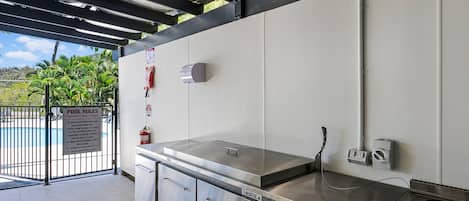 Een keuken met roestvrijstalen apparatuur, een wit aanrechtblad en een bord met zwembadregels zichtbaar door een open poort.