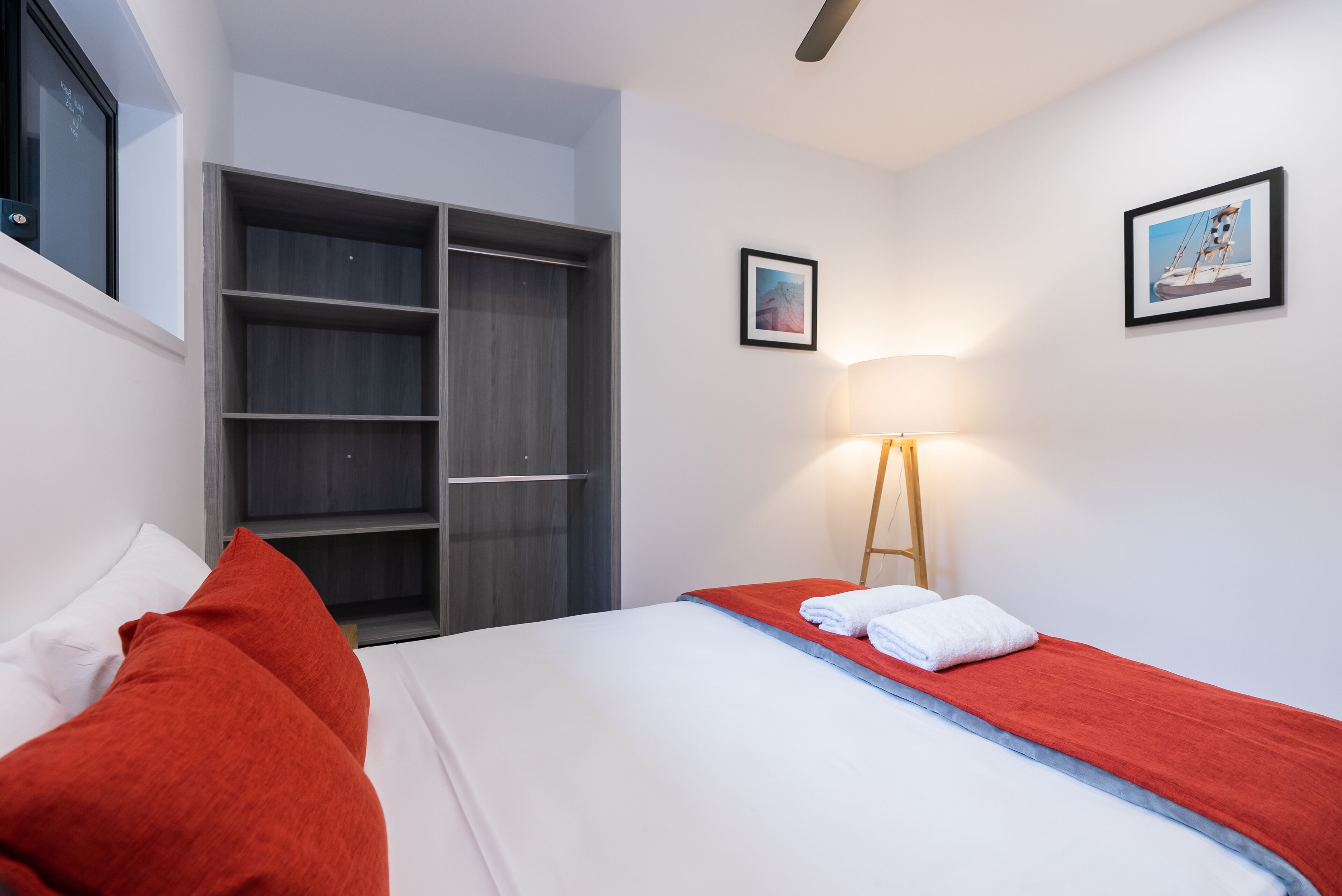 Una habitación de hotel con una cama, ropa de cama roja y blanca, un armario, una mesita de noche con una lámpara y dos cuadros en la pared.