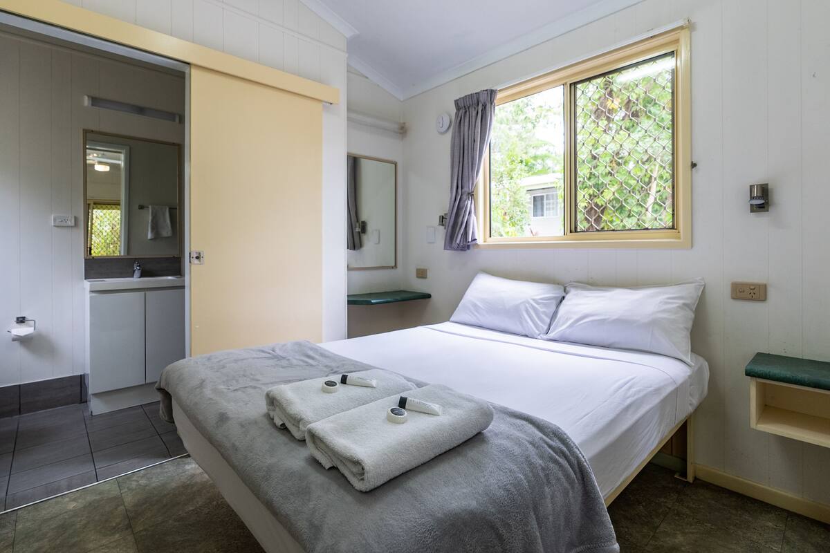 Une chambre d’hôtel avec un lit, une salle de bain, une fenêtre donnant sur un espace verdoyant et un miroir.