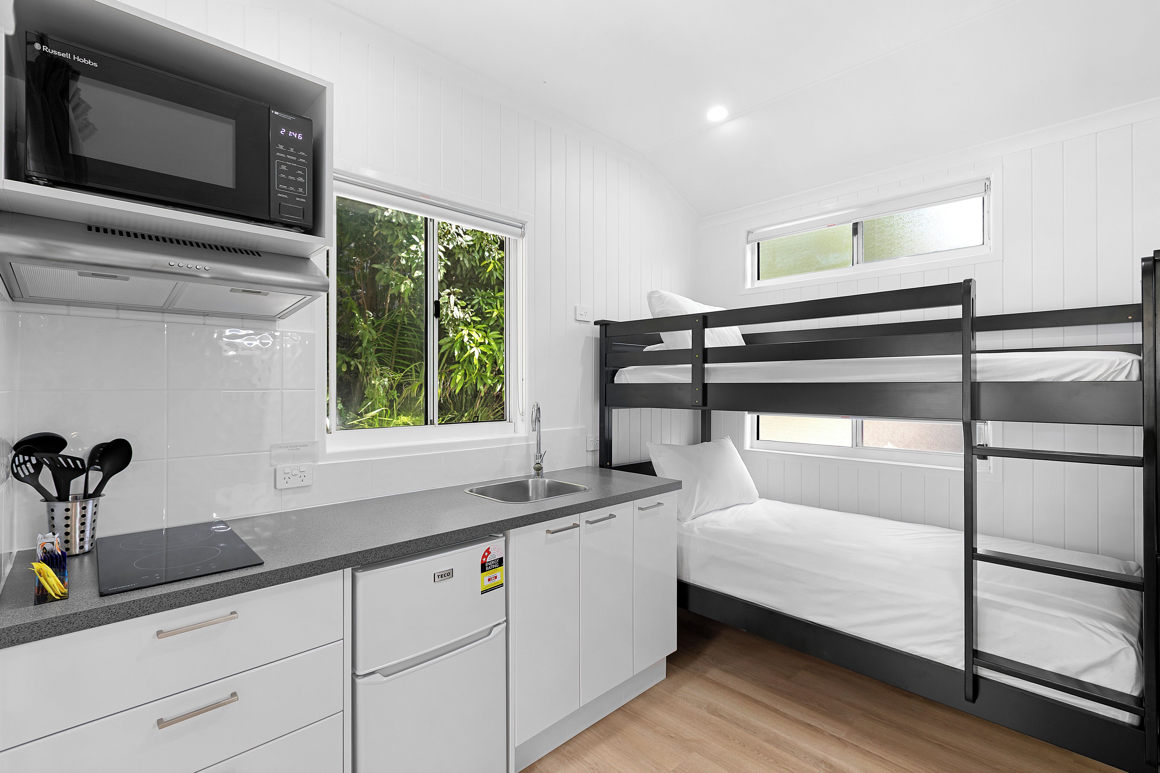 A compact kitchenette with a microwave, sink, and cabinets. There is a bunk bed with a window above it.