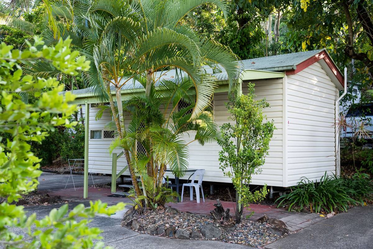 A small, single-story house with a green roof and white siding, surrounded by lush greenery and a small patio area with a chair and table.