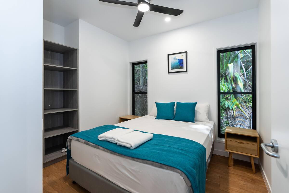 Une chambre moderne comprenant un lit, un ventilateur de plafond, une table de chevet, une fenêtre donnant sur un espace verdoyant et une bibliothèque intégrée.
