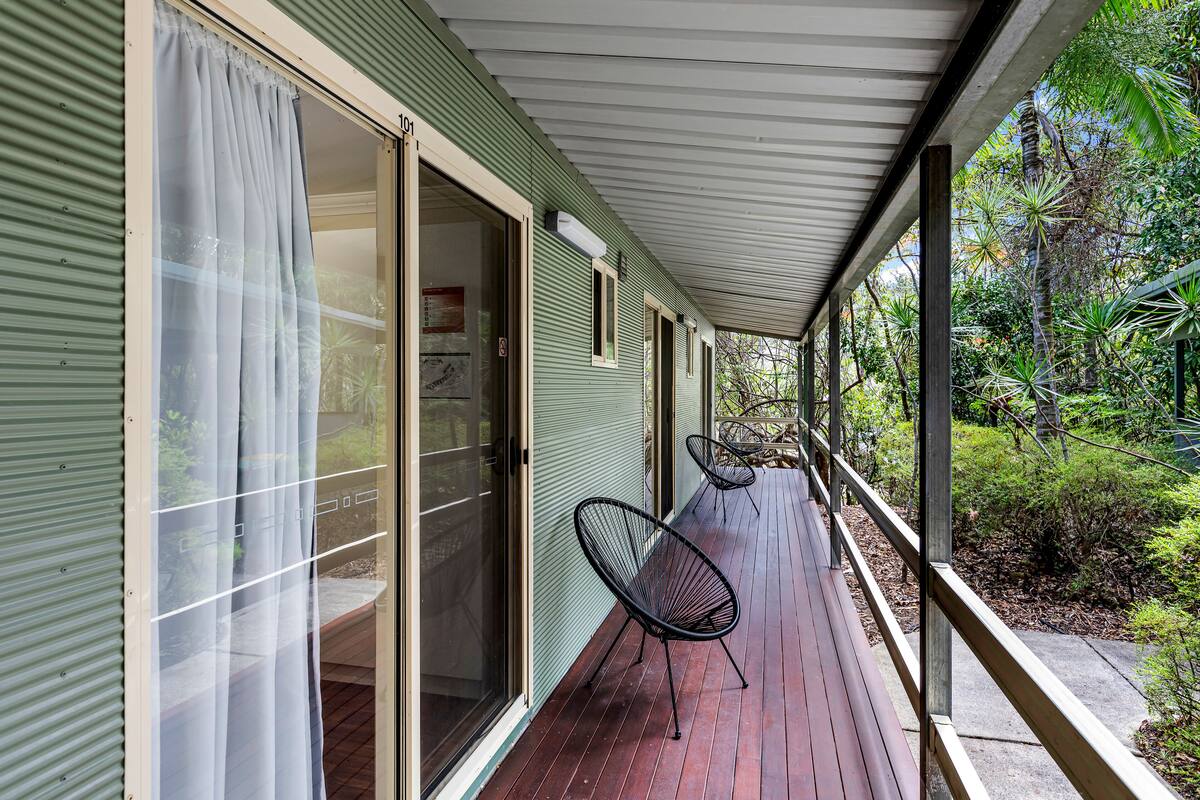 Une véranda couverte avec une terrasse en bois, des portes coulissantes en verre et deux chaises noires en fil de fer.