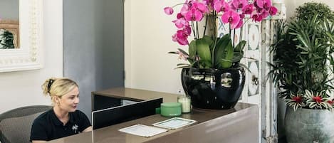 Un mostrador de recepción con una orquídea grande en maceta, un cuadro enmarcado y una planta grande a la derecha.