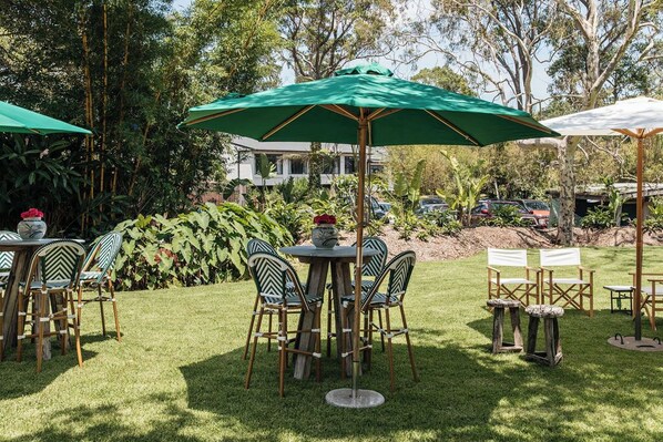 An outdoor patio area with round tables and chairs, green umbrellas, and a lush garden backdrop.