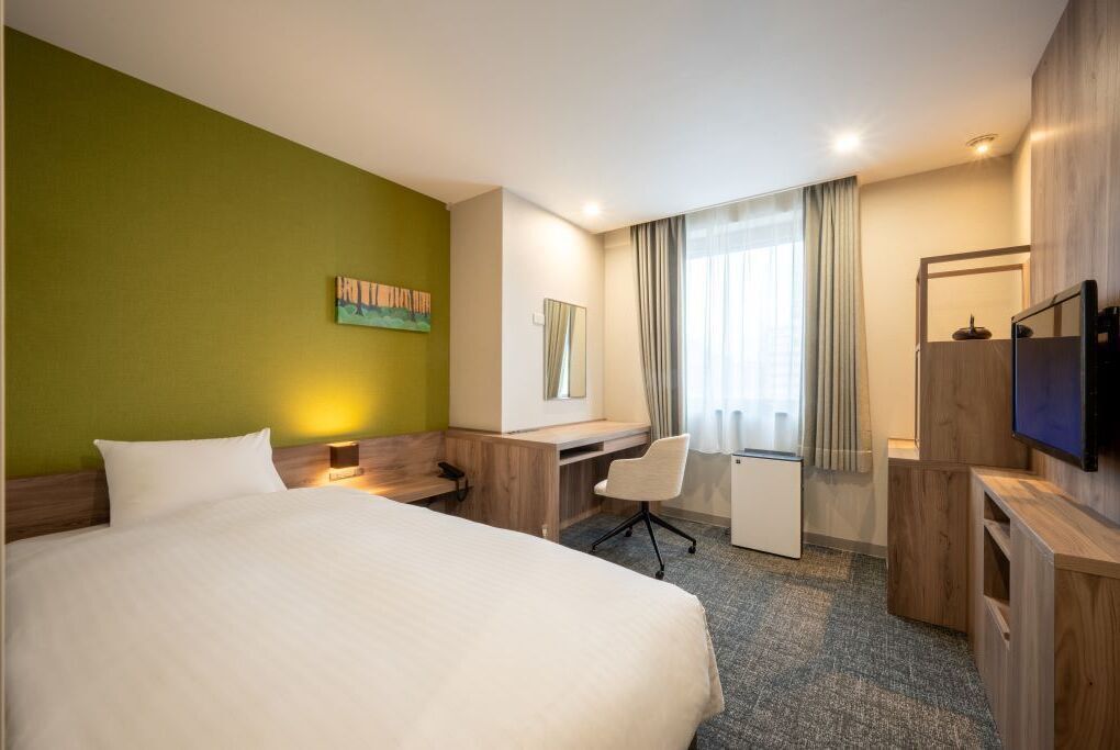 Habitación de hotel con cama, escritorio, silla, televisión y ventana con cortinas.