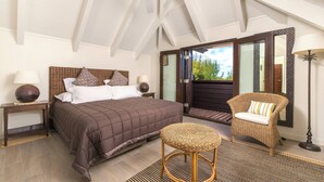 A bedroom with a large bed, two nightstands, a wicker chair, a small round table, and a view of the outdoors through a glass door. - Te Vakaroa Villas (Rarotonga)