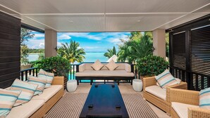 A sunlit outdoor seating area with a sofa, two armchairs, and a coffee table, overlooking a scenic view of the ocean and palm trees. - Te Vakaroa Villas (Rarotonga)