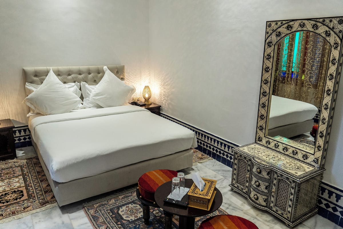 A neatly made bed with white linens, a bedside lamp, a small table with a remote control, a tissue box, and a decorative mirror.