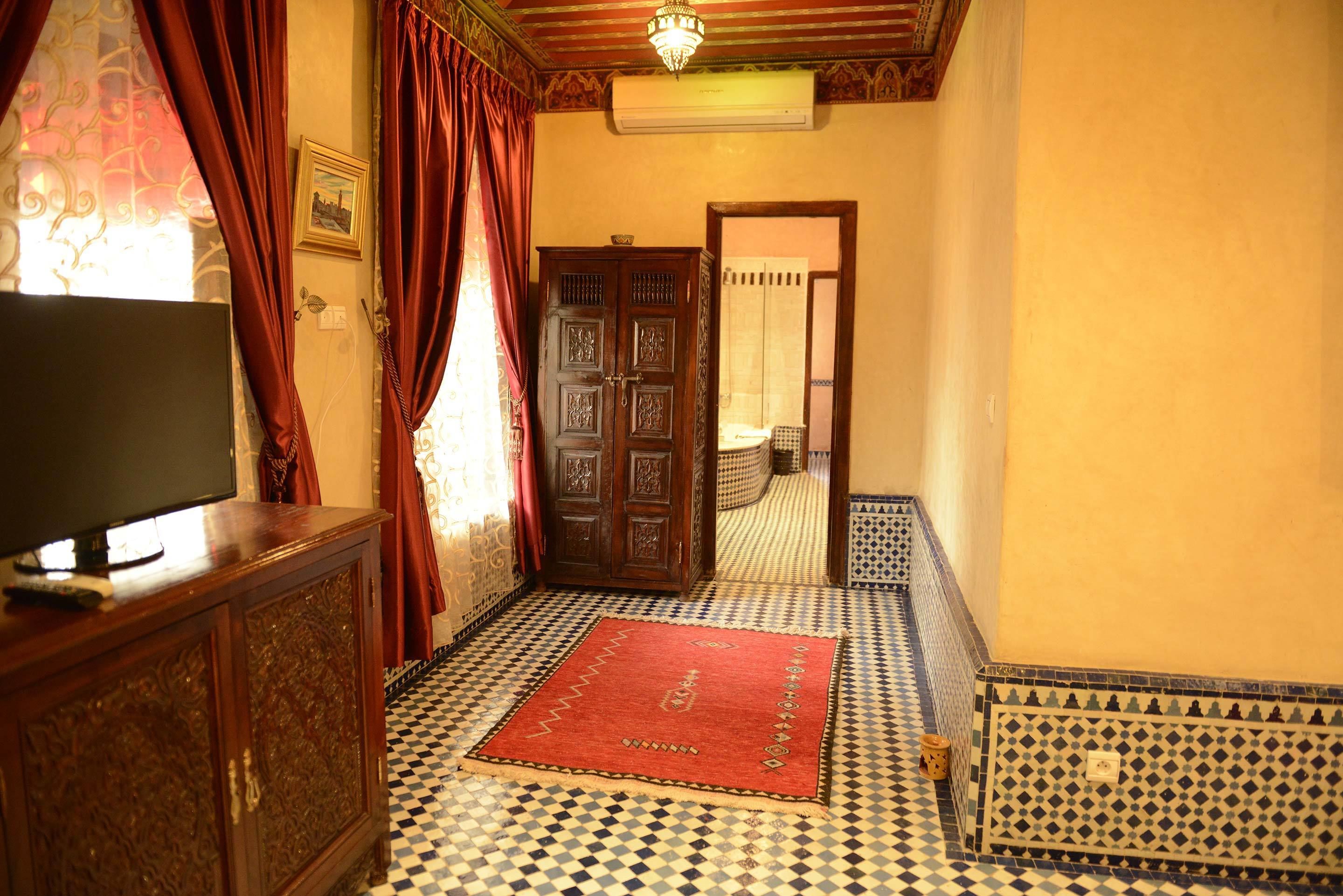 Un pasillo con piso a cuadros, un tapete rojo, un mueble de madera con un televisor y una puerta con intrincados grabados.