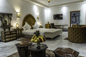 Quarto de hotel espaçoso com uma cama grande, televisão, área de refeições com uma cesta de frutas e decoração tradicional.