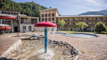 A fountain with a red top, surrounded by a stone border, in front of a building with multiple balconies and a pool.