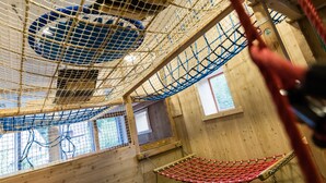 Une structure de jeu en bois, dotée de zones d’escalade avec des filets et d’une balançoire rouge.