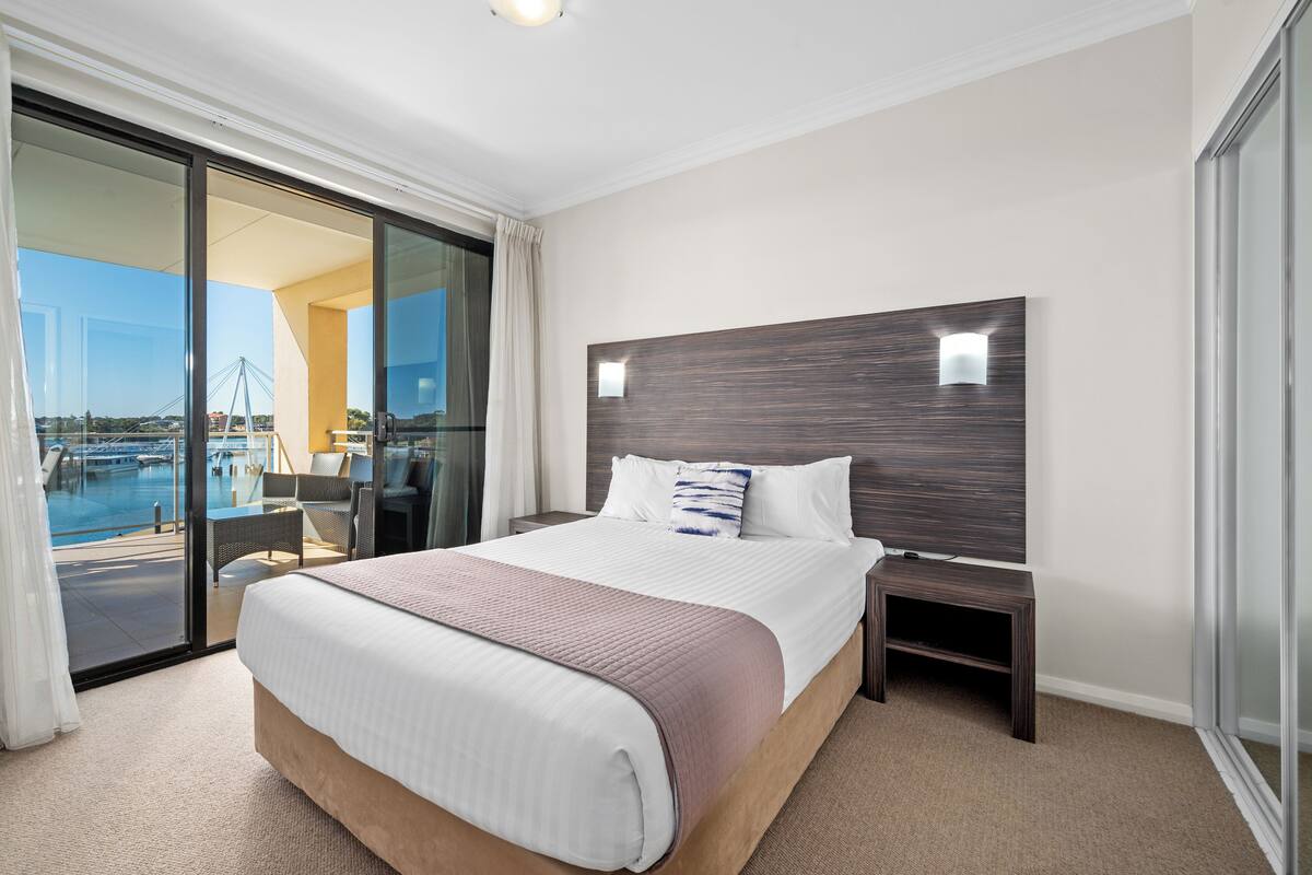 A hotel room with a large bed, a nightstand, a glass door leading to a balcony with a view of a bridge and water.
