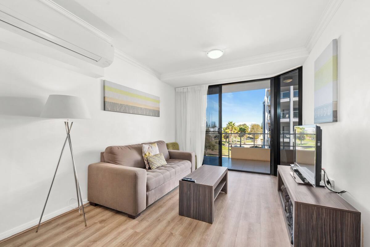 A modern living room with a sofa, coffee table, and TV. It features a large window with a balcony view.