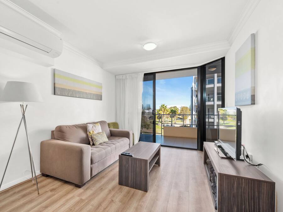 A modern living room with a sofa, coffee table, and TV. It features a large window with a balcony view.