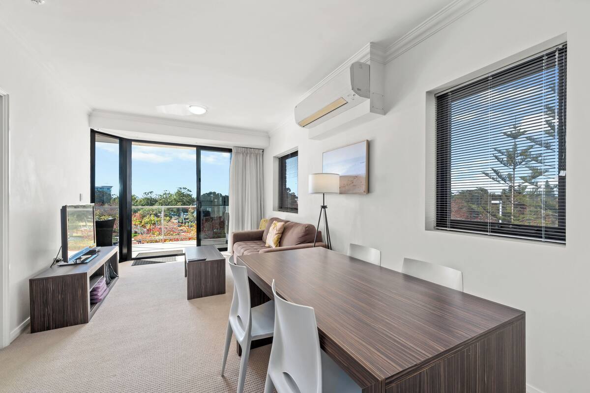 A modern room with a large dining table, white chairs, a sofa, and a TV on a stand.