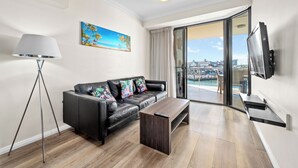 A modern living room with a black leather sofa, a wooden coffee table, and a floor lamp. A painting of a tropical scene hangs on the wall, and a TV is mounted on the wall. - Dolphin Quay Apartments (Mandurah)