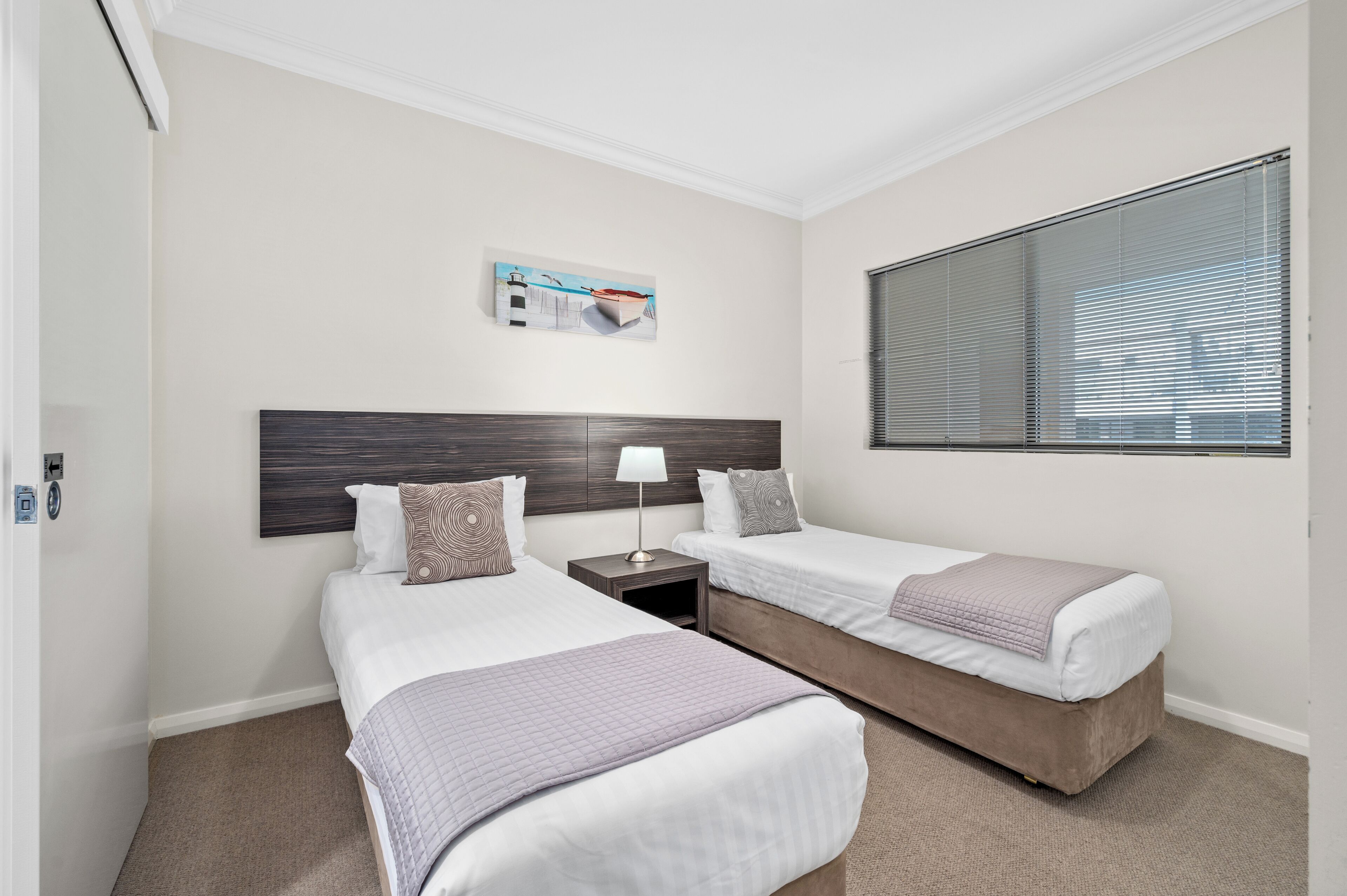 A hotel room with two single beds, a nightstand with a lamp, a painting on the wall, and a window with blinds.