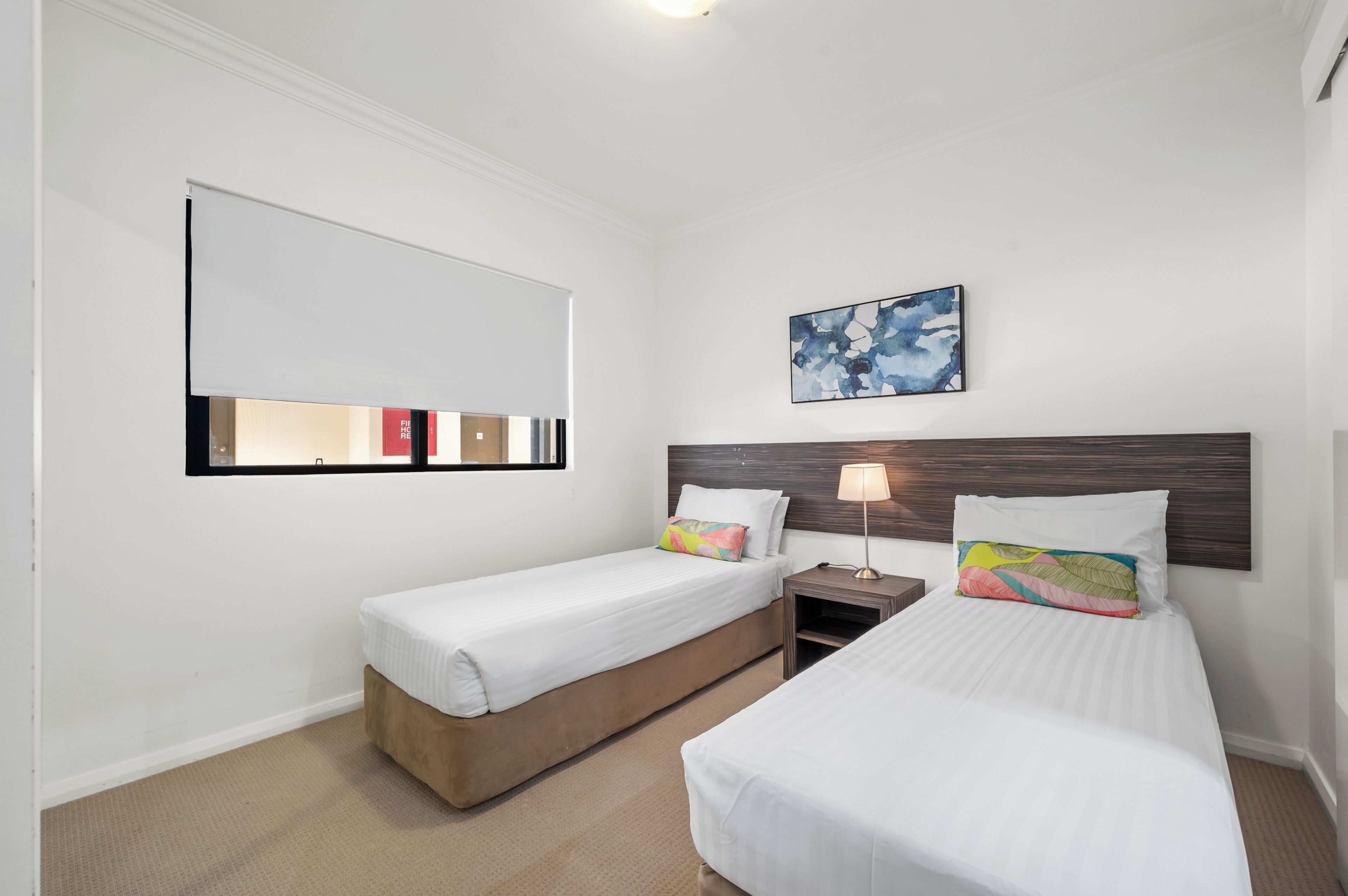 A hotel room with two single beds, a nightstand with a lamp, a large window with a view, and a wall-mounted artwork.