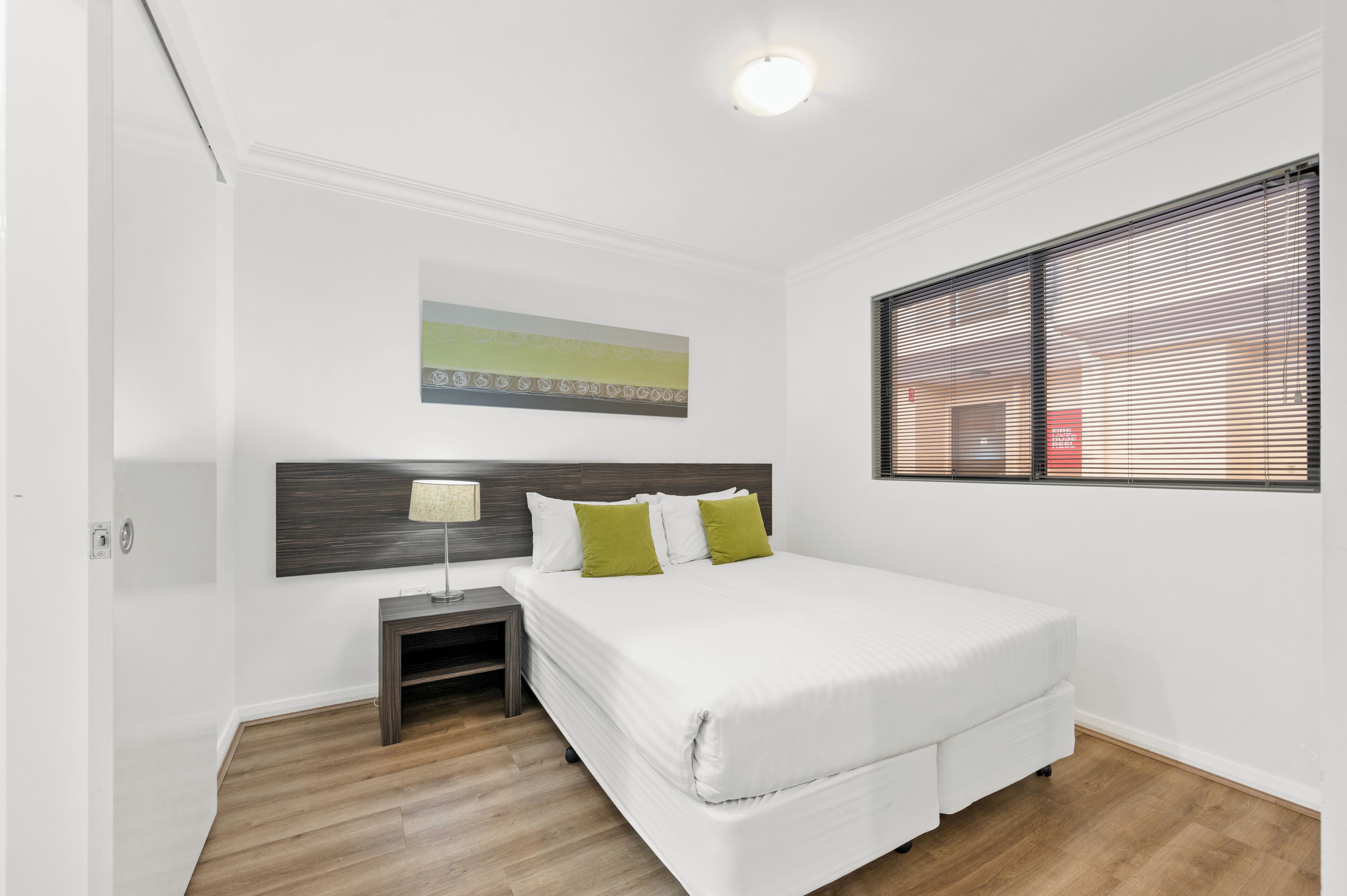Un dormitorio con cama, mesita de noche, lámpara, un cuadro y ventana con persianas.