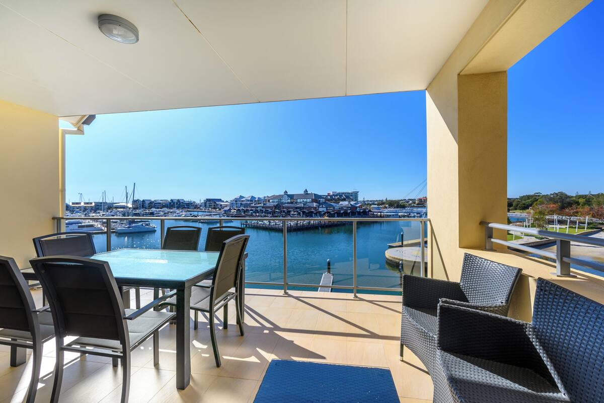 A balcony with a view of a marina, featuring a table and chairs, a railing, and a clear sky.