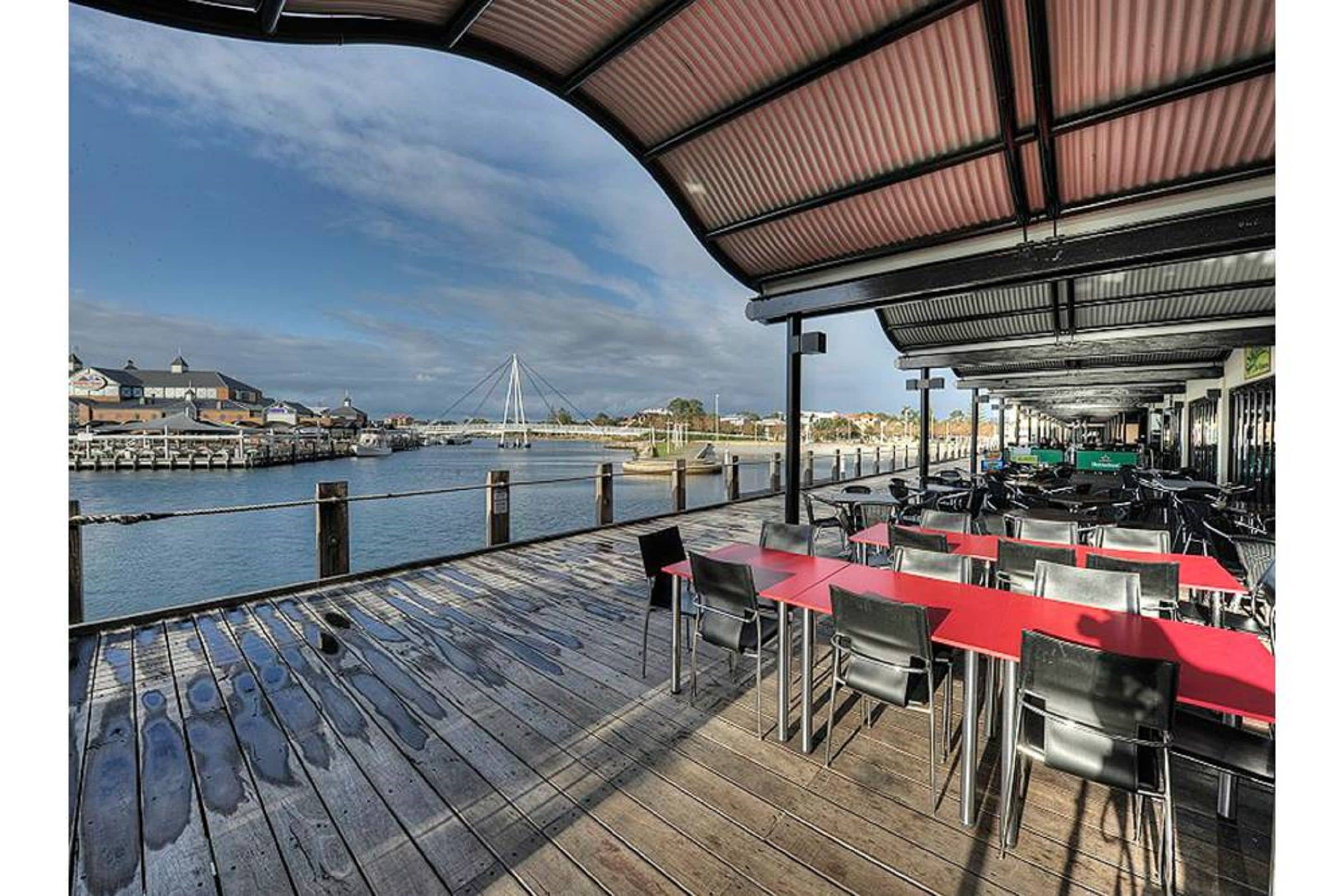 An outdoor dining area with red tables and chairs, overlooking a river with a bridge in the background.