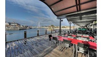 An outdoor dining area with red tables and chairs, overlooking a river with a bridge in the background.