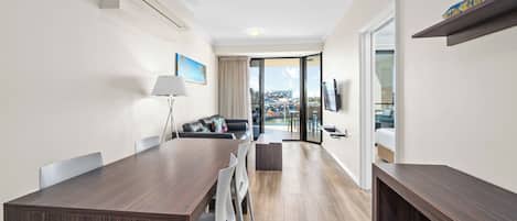 A modern hotel room with a dining area, a sofa, a TV, and a balcony with a city view.