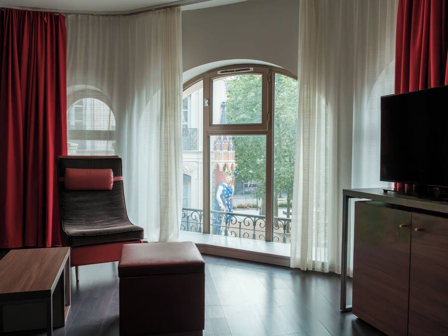 Une chambre moderne avec une grande fenêtre, un fauteuil rouge, un banc en bois et un téléviseur posé sur un meuble.