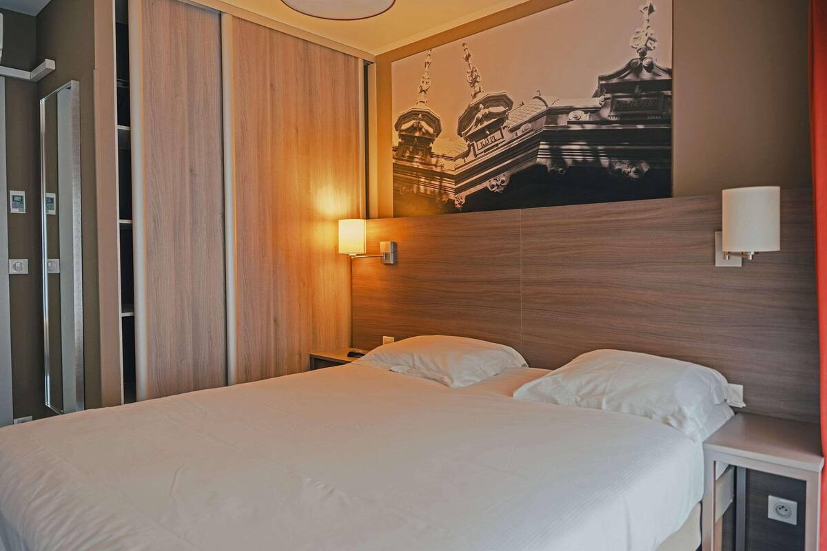 Habitación de hotel con una cama, una mesita de noche, una lámpara de pared y una gran obra de arte mural que representa un edificio.