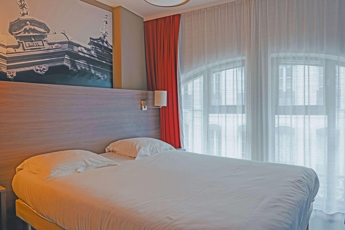Habitación de hotel con cama, cabecero, lámpara de pared y ventana con cortinas de gasa.