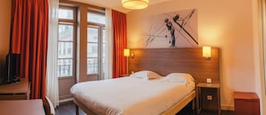 Une chambre d’hôtel avec un lit, une table de chevet, une lampe, un tableau et une fenêtre avec des rideaux.