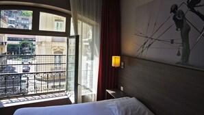 A hotel room with a bed, a window with a view of buildings and a street, a statue on the wall, and a lamp.